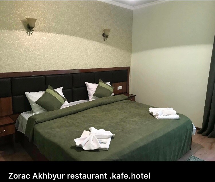 A well-appointed sleeping area features a spacious bed dressed in green linens, complemented by white pillows and towels. Soft lighting is provided by wall-mounted fixtures. The room's light green walls create a calming atmosphere, with natural light entering through a nearby window dressed in dark curtains.