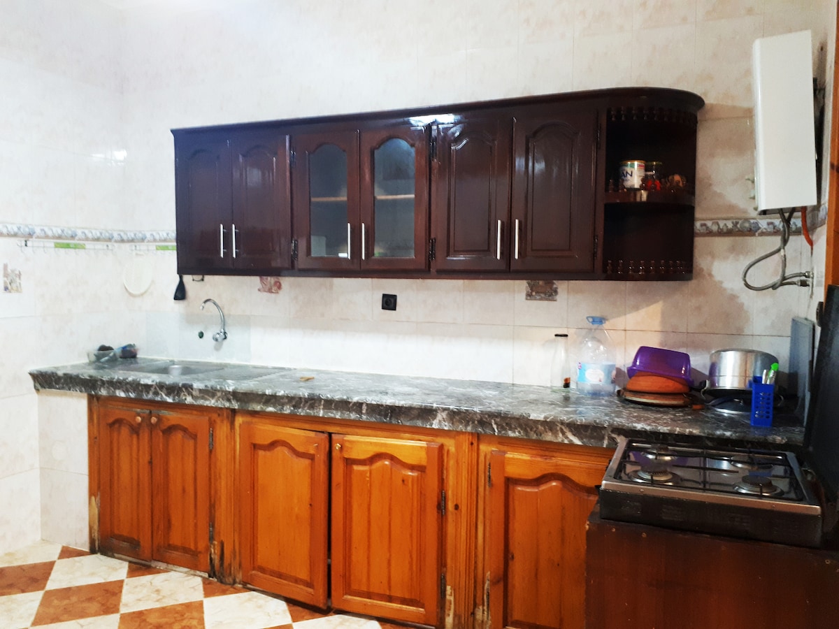 A kitchen is showcased with wooden cabinetry and a dark marble countertop. The cabinets feature glass-paneled doors. A gas stove is present, along with various kitchen essentials, including a sink and appliances, all arranged in a functional layout.