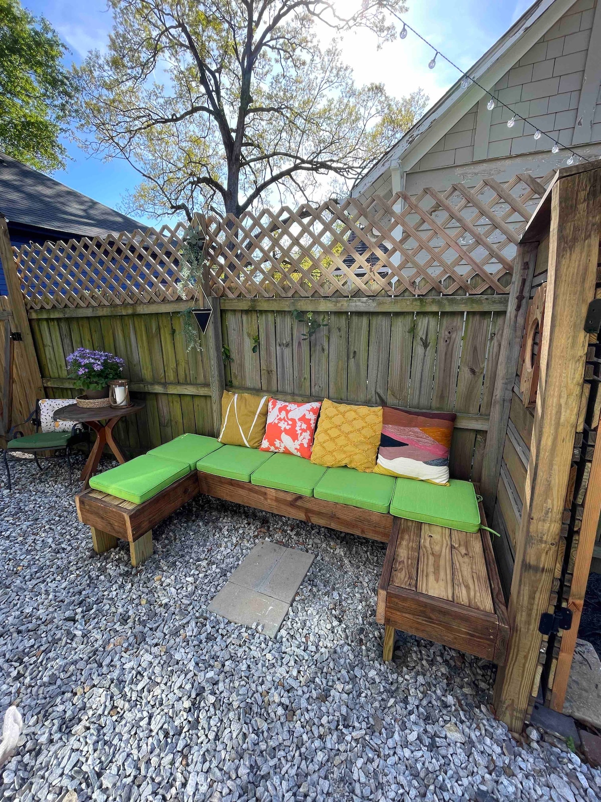 An outdoor seating area is showcased, featuring a wooden bench adorned with colorful cushions in shades of yellow, green, and red. A small wooden table is positioned nearby, surrounded by gravel, with greenery visible beyond the wooden lattice. Natural light filters through, creating a welcoming space.