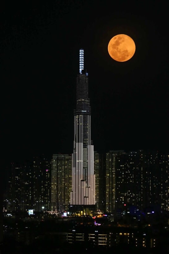 A towering skyscraper is illuminated against a night sky, with its unique architectural features clearly displayed. A large, orange moon hovers above, contrasting against the bright city lights of the buildings in the background, creating a striking urban landscape.