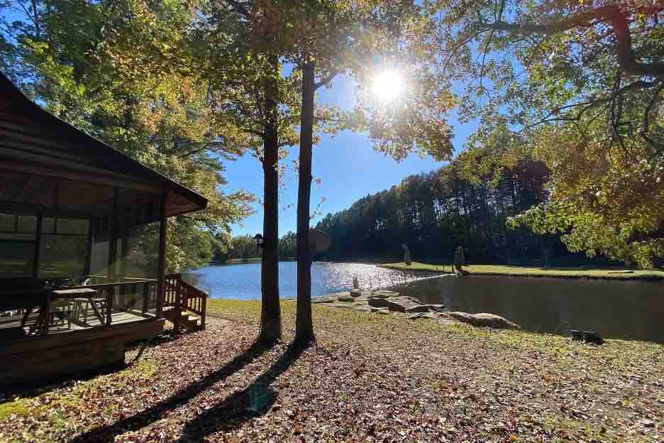 Sunlight filters through the trees, casting a warm glow over the serene lake. A cabin sits partially visible, with a seating area on the deck facing the water. The landscape is framed by lush greenery and rocky accents, creating a tranquil natural setting.