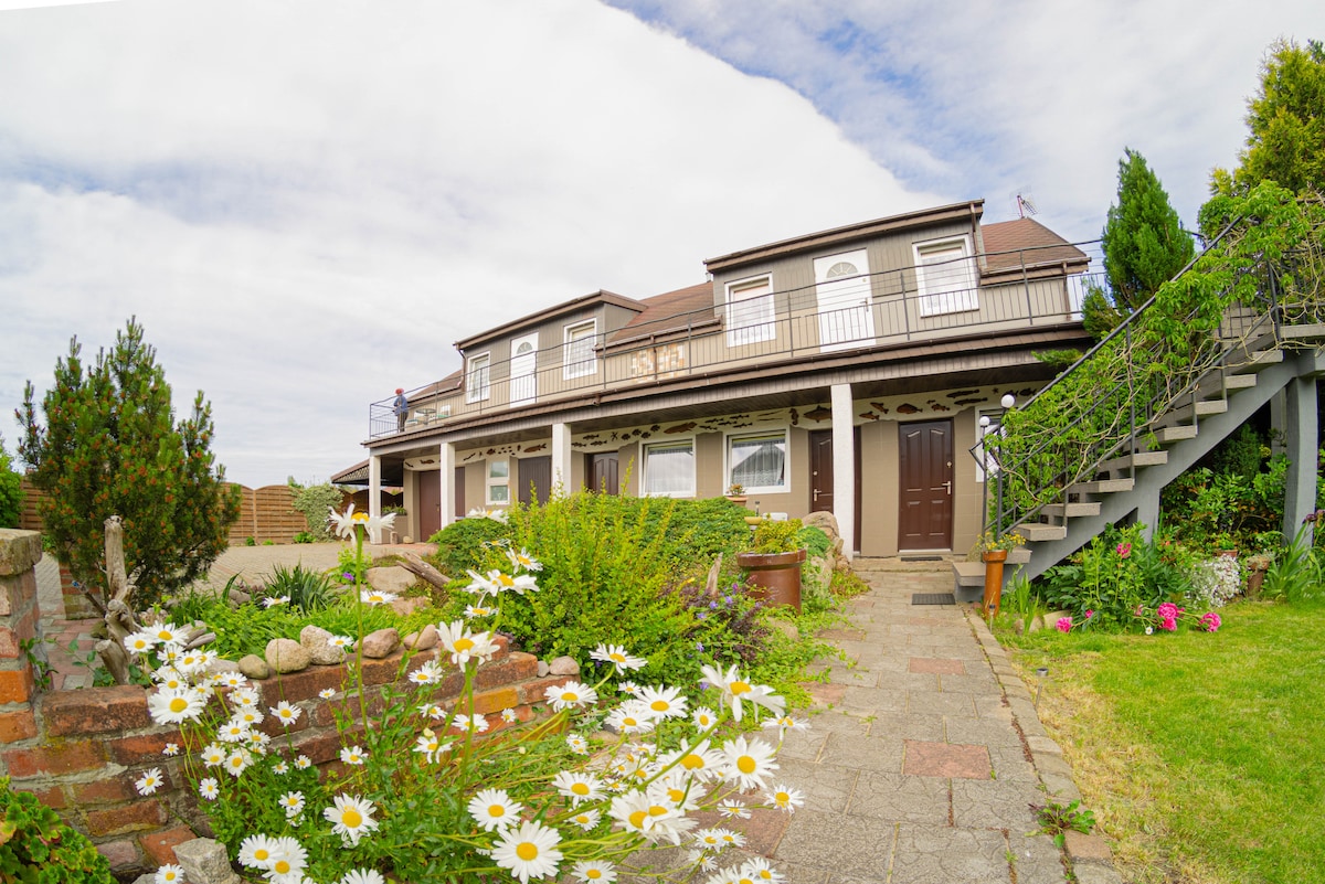The exterior of the building exhibits a multi-level structure with a welcoming entrance. A well-maintained garden with blooming daisies and lush greenery surrounds the entrance, enhancing the overall charm of the property. A staircase leads to the upper level, flanked by potted plants.