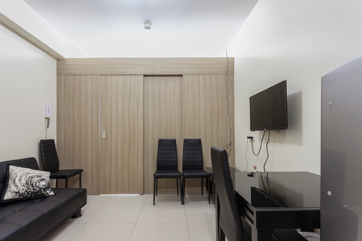 A spacious living area features a black sofa bed and two sleek black chairs. A dark dining table is positioned in front of a wall-mounted television. The room is complemented by light-colored walls and gray tiles, creating a modern and functional atmosphere.