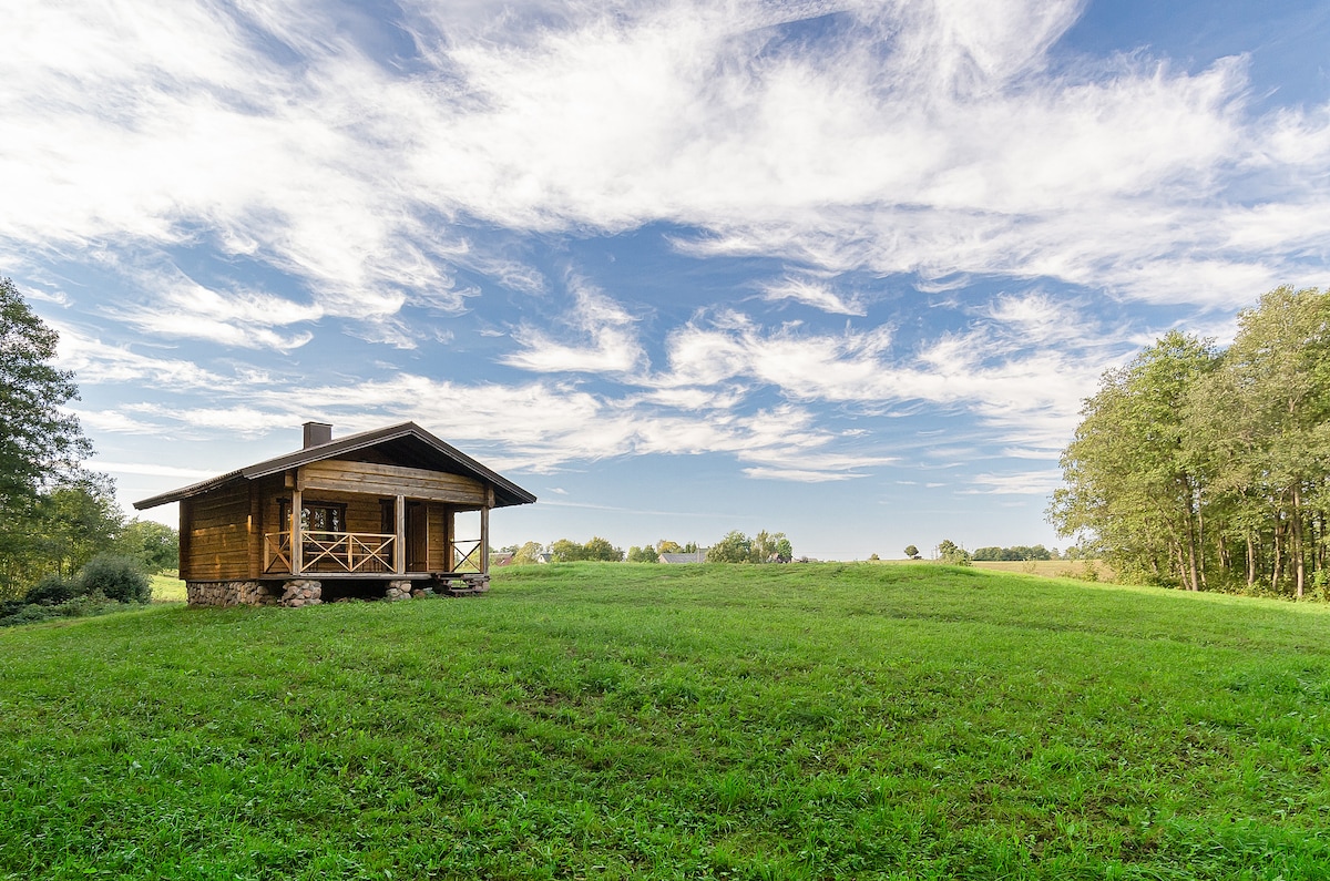 A wooden cabin is positioned amidst an expansive green landscape, with a clear blue sky overhead adorned with scattered clouds. Lush grass surrounds the cabin, providing a serene environment enhanced by nearby trees along the perimeter.