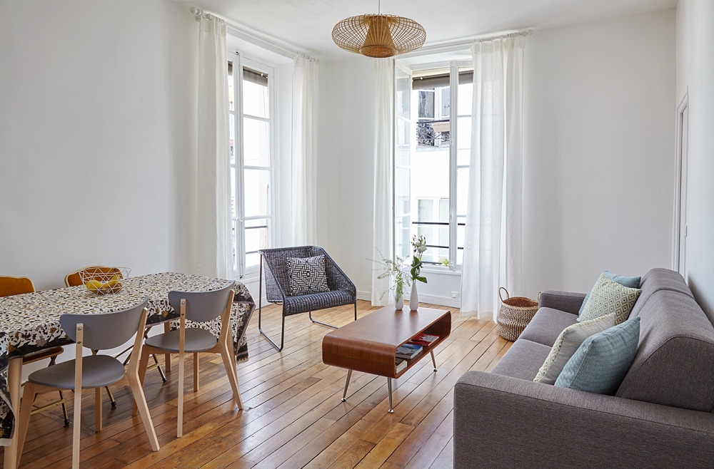A bright living area is furnished with a gray sofa, a wooden coffee table, and a patterned rug. A dining table with several chairs is visible, complemented by natural light from the large windows covered with sheer white curtains.