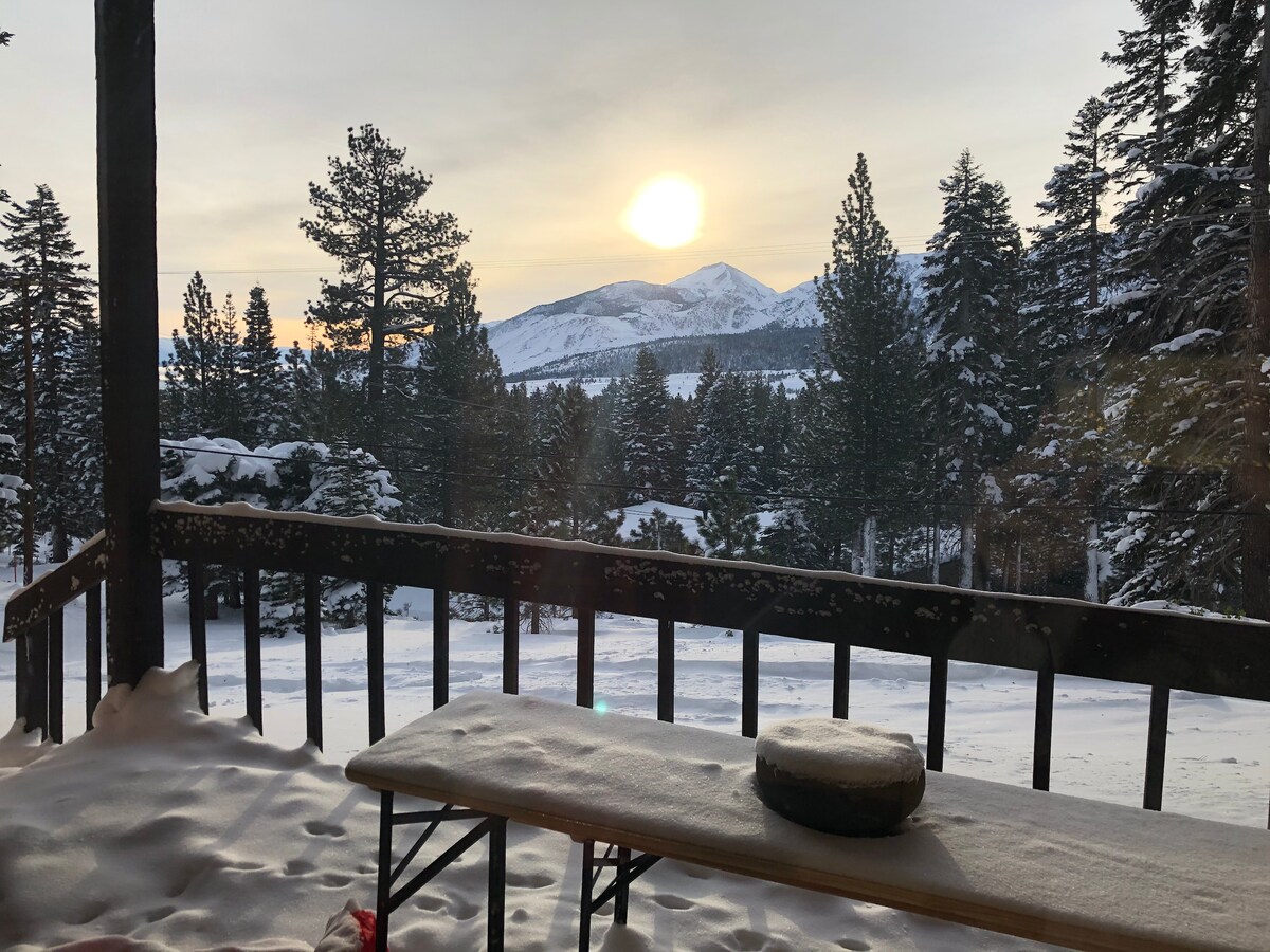A snowy deck is viewed, framed by wooden railings, with a round table set on its surface. The sun sets behind distant mountains and trees, casting a warm glow over the winter scene, while snow covers the ground and branches.