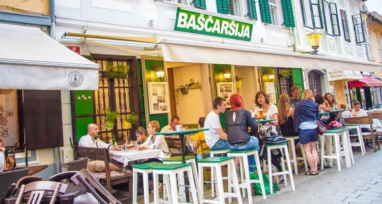 An outdoor dining area is shown with several tables occupied by guests enjoying meals. Stools with green and white accents surround the tables. The restaurant's name, 'Baščarija,' is displayed prominently above the entrance, complemented by green shutters on the windows which add to the inviting atmosphere.