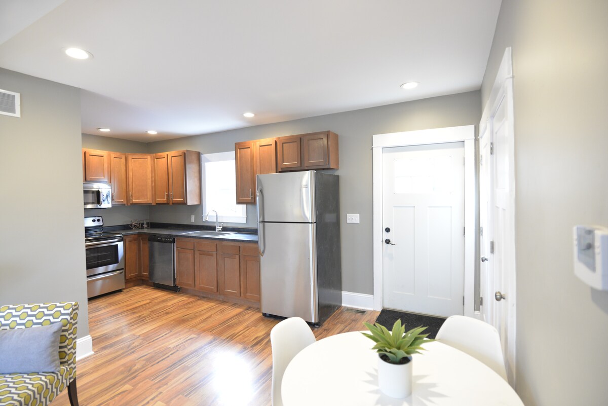 The kitchen area showcases wooden cabinetry and modern stainless steel appliances, including a refrigerator and stove. A round dining table with white chairs is positioned near the entrance, with bright lighting enhancing the space's clean, welcoming look.