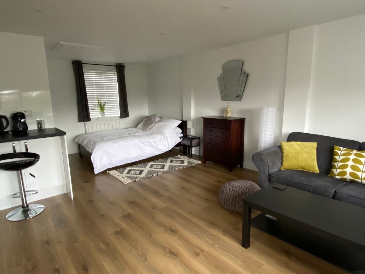 A spacious studio is presented, featuring a double bed with crisp white bedding and a dark wood dresser. A gray sofa, accented by a yellow pillow, sits across from a small coffee table. Natural light enters through window blinds, illuminating the warm wood flooring.
