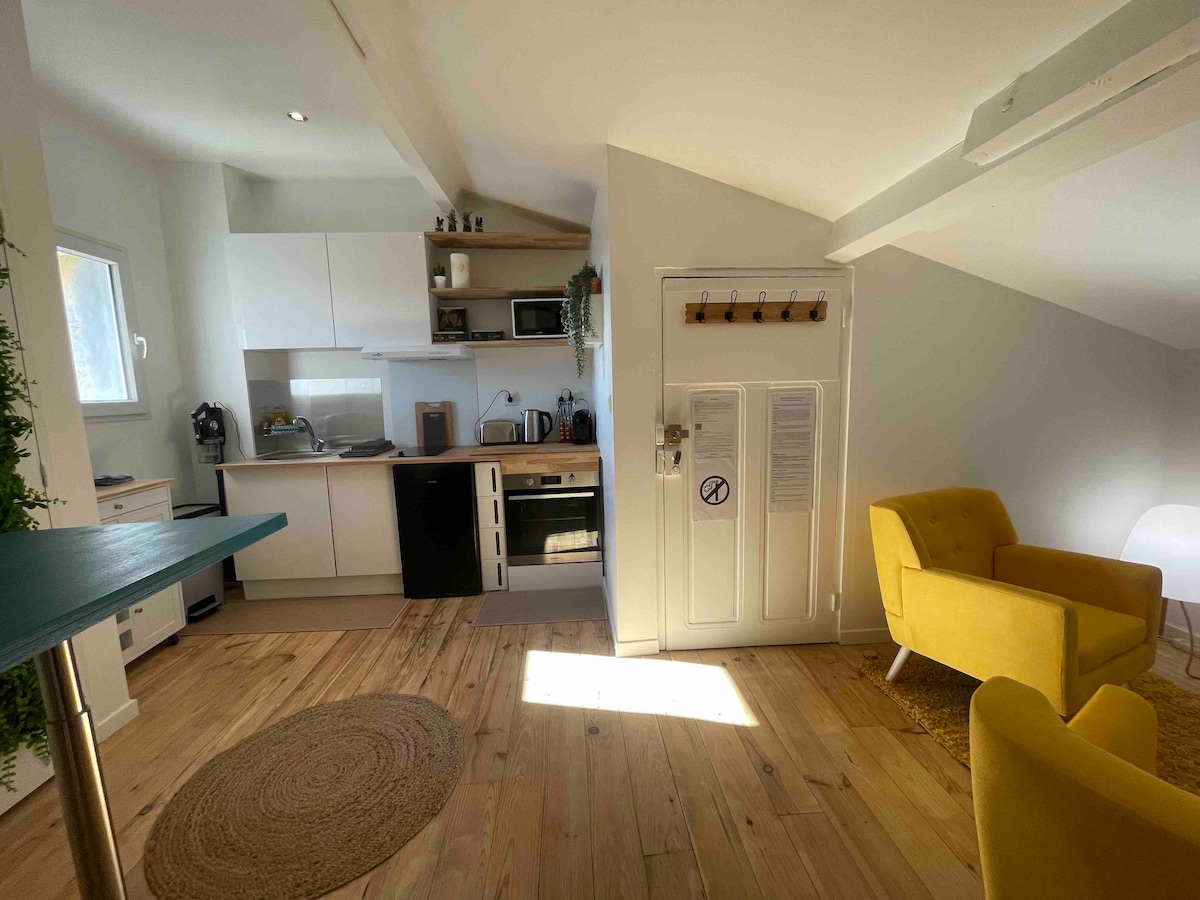 The open kitchen area features modern appliances, including a stove and microwave, alongside an adjoining living space with a bright yellow armchair. Natural light floods the area through a window, highlighting the light wood flooring and a round area rug placed on the floor.