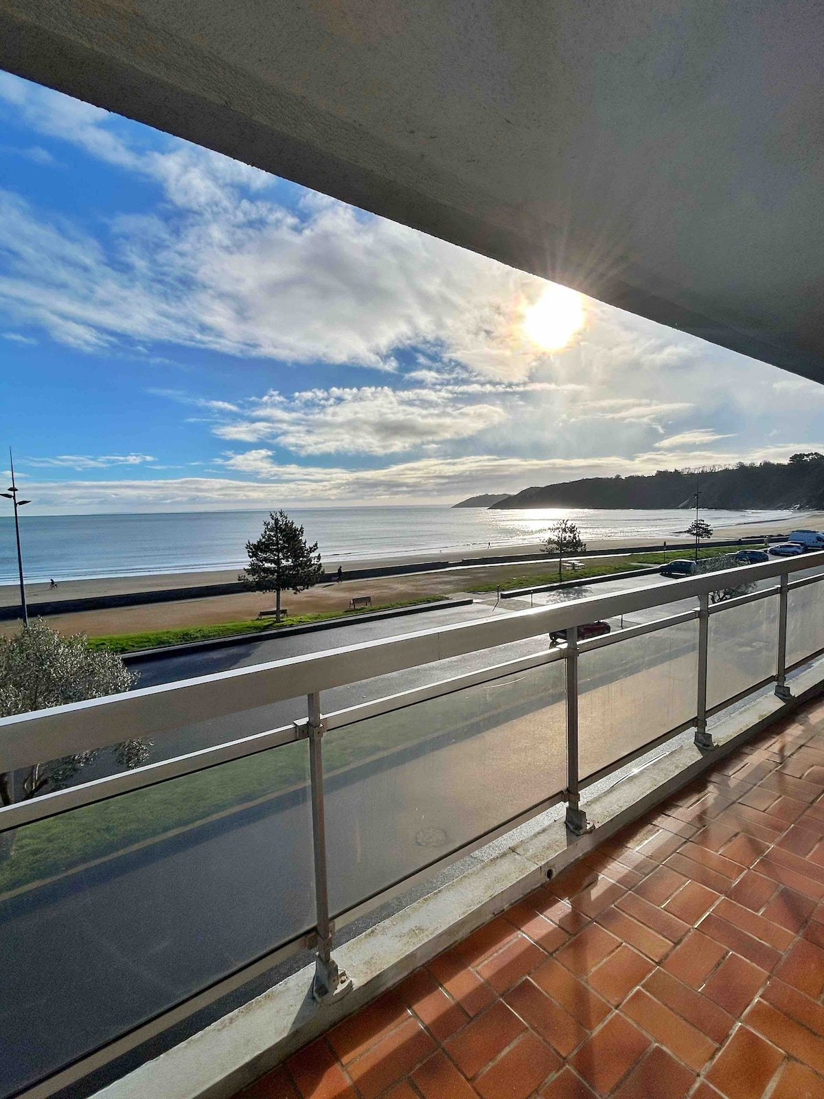 A balcony offers a scenic view of the beach, with sunlight reflecting on the water. The coastline is lined with trees, and a promenade can be seen in the foreground. The sky features a mix of clouds and blue, enhancing the coastal ambiance.
