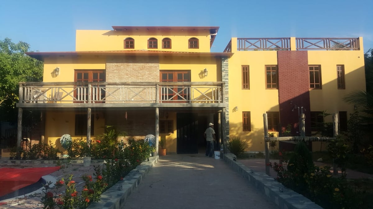 A vibrant two-story structure features a yellow exterior with stone accents. A spacious balcony is supported by wooden railings, overlooking a well-maintained garden with colorful flowers. The pathway leading to the entrance is framed by greenery, enhancing the welcoming ambiance.