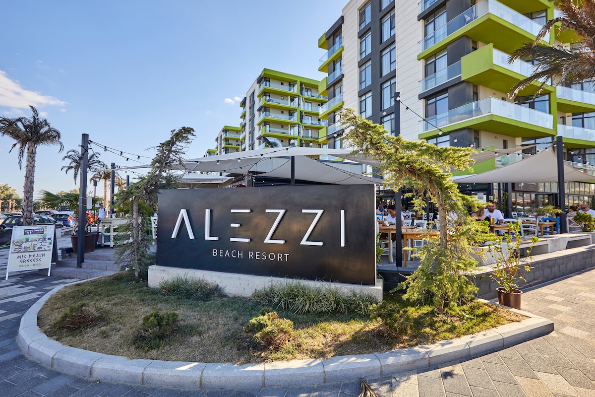 The entrance of Alezzi Beach Resort is prominently displayed, featuring the bold sign with 'Alezzi Beach Resort' in white on a dark background. Surrounding greenery and outdoor dining areas are visible, highlighting the lively atmosphere of the resort with modern buildings in the background.