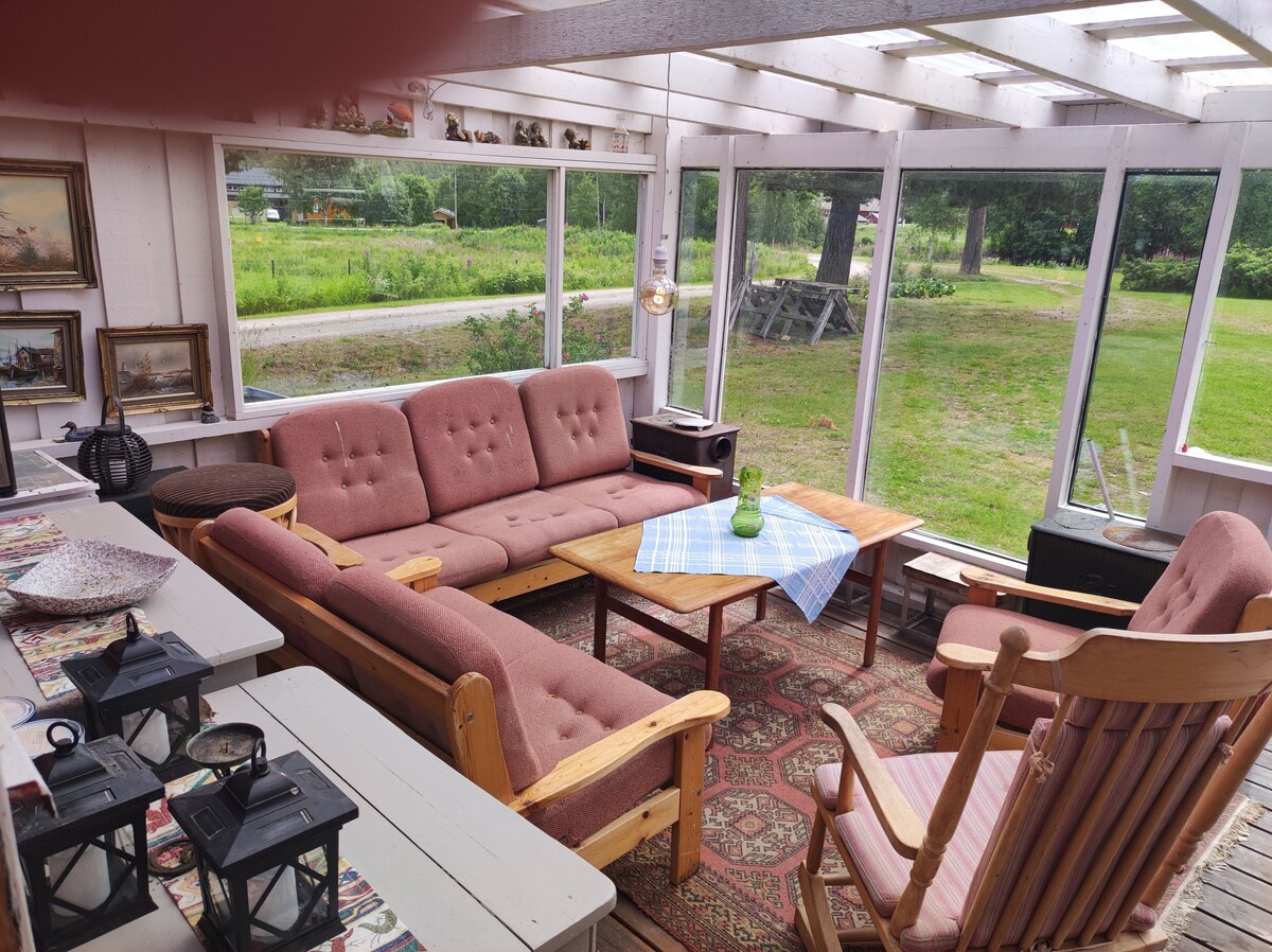 A sunlit enclosed veranda features a wooden table at the center, surrounded by a sofa and armchair. Large windows offer views of the surrounding greenery, while decorative rugs add warmth to the space. Lanterns are placed on a nearby surface, enhancing the inviting atmosphere.