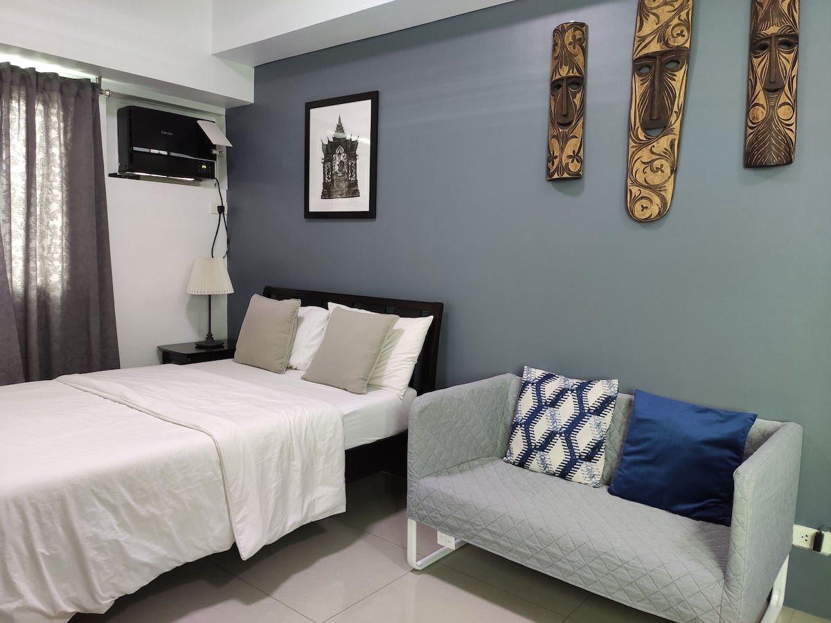 A cozy studio unit features a spacious bed with soft linens and decorative pillows, complemented by a stylish sofa. Textured wall art adds character to the room, while natural light filters through the window drapes, creating a comfortable ambiance.