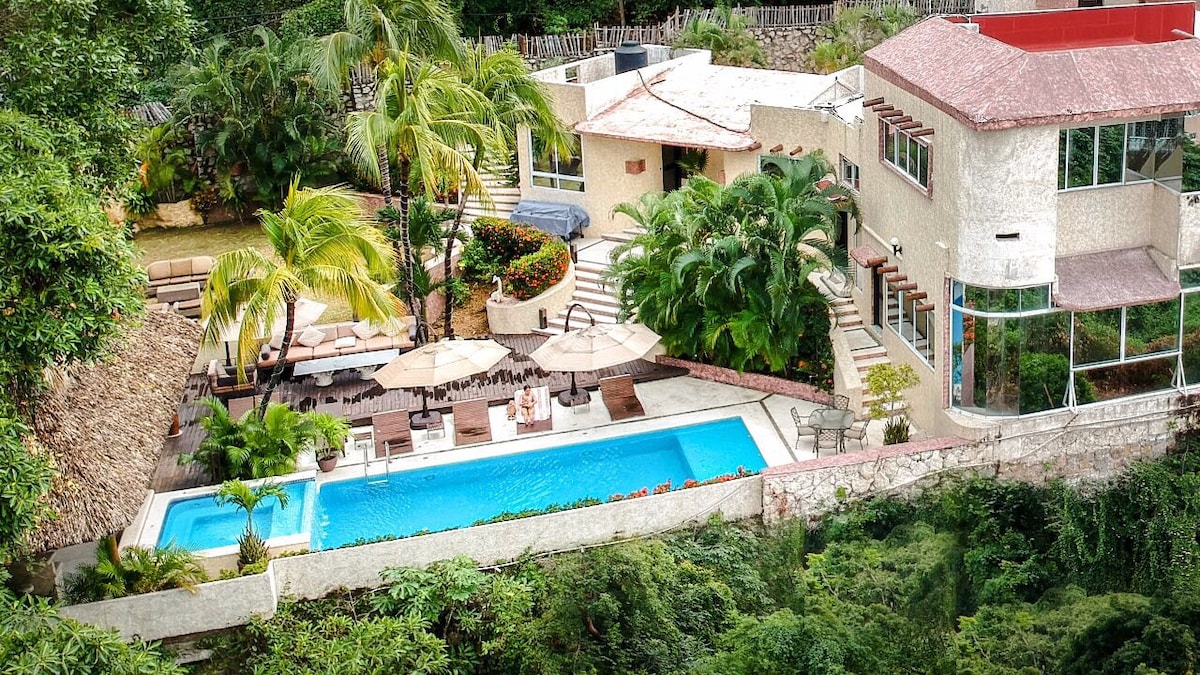 An aerial view showcases a landscaped outdoor area featuring a shimmering pool surrounded by lounge chairs and umbrellas. Lush palm trees and tropical plants enhance the space, while well-placed steps lead to various levels, offering an inviting atmosphere for relaxation and leisure.