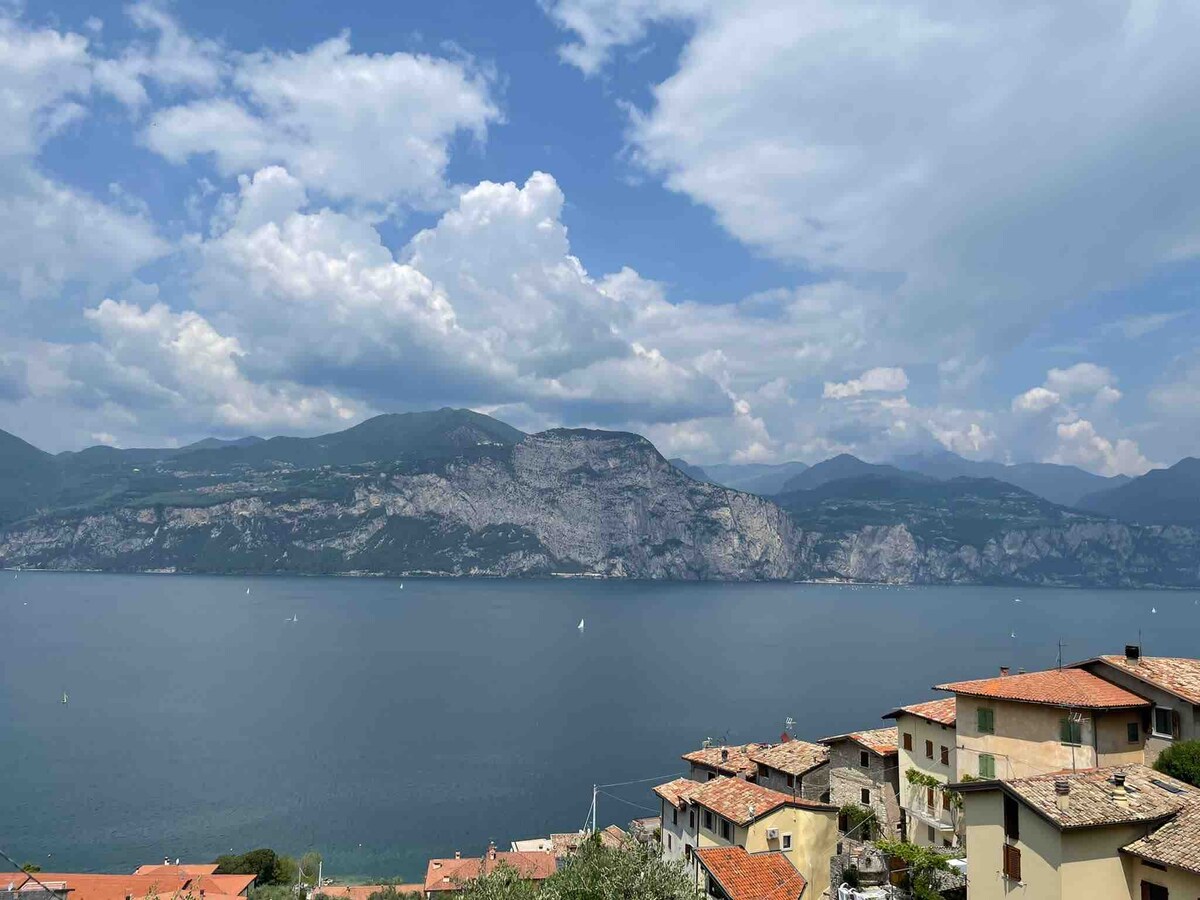 A panoramic view of Lake Garda is captured, with distant mountains standing majestically against a partly cloudy sky. The calm waters reflect the landscape, while the charming rooftops of nearby buildings frame the foreground, showcasing the serene beauty of this lakeside setting.