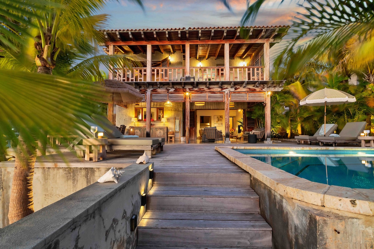 The villa is enveloped by lush palm trees and features a spacious wooden deck leading to a large swimming pool. The two-story structure accommodates a terrace with seating, with outdoor loungers positioned for relaxation. Soft lighting highlights the inviting ambiance during the evening hours.