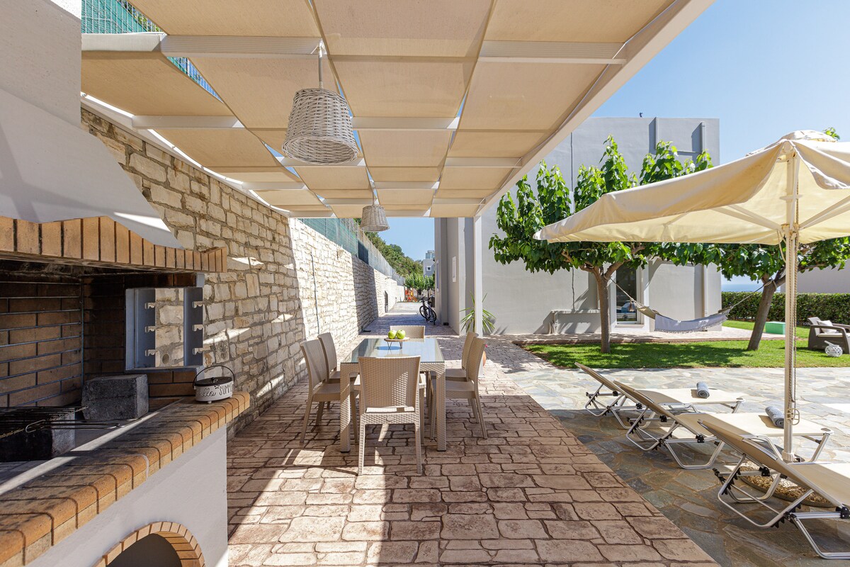 An outdoor dining area features a shaded table and chairs, accompanied by a built-in barbecue. Sun loungers are arranged under large umbrellas, providing a comfortable space for relaxation. The surrounding garden is visible, enhancing the outdoor experience with greenery.