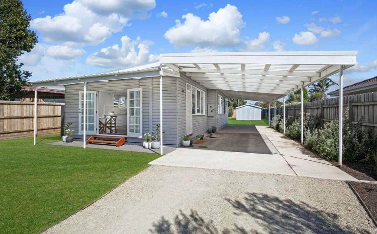 A coastal retreat is showcased, featuring a light grey exterior with large windows and a spacious carport. The grassy yard is well-maintained, while the walkway leads to a seating area on the porch, which is surrounded by blooming shrubs and trees.