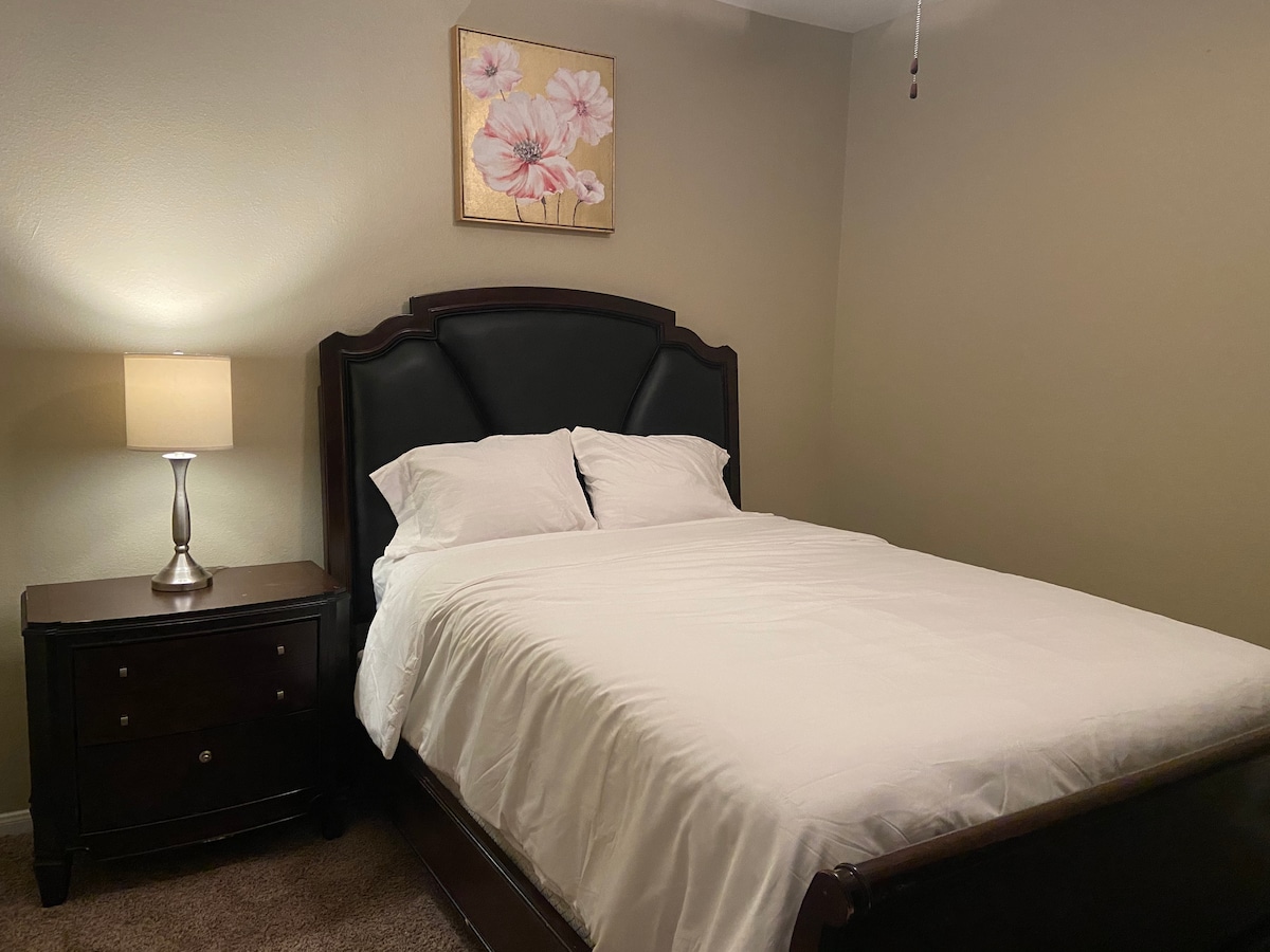 A cozy bedroom features a spacious bed with white linens and two decorative pillows. A nightstand with a lamp stands beside the bed, contributing to the warm lighting. A floral artwork piece is positioned above the bed, adding a subtle touch to the room's decor.