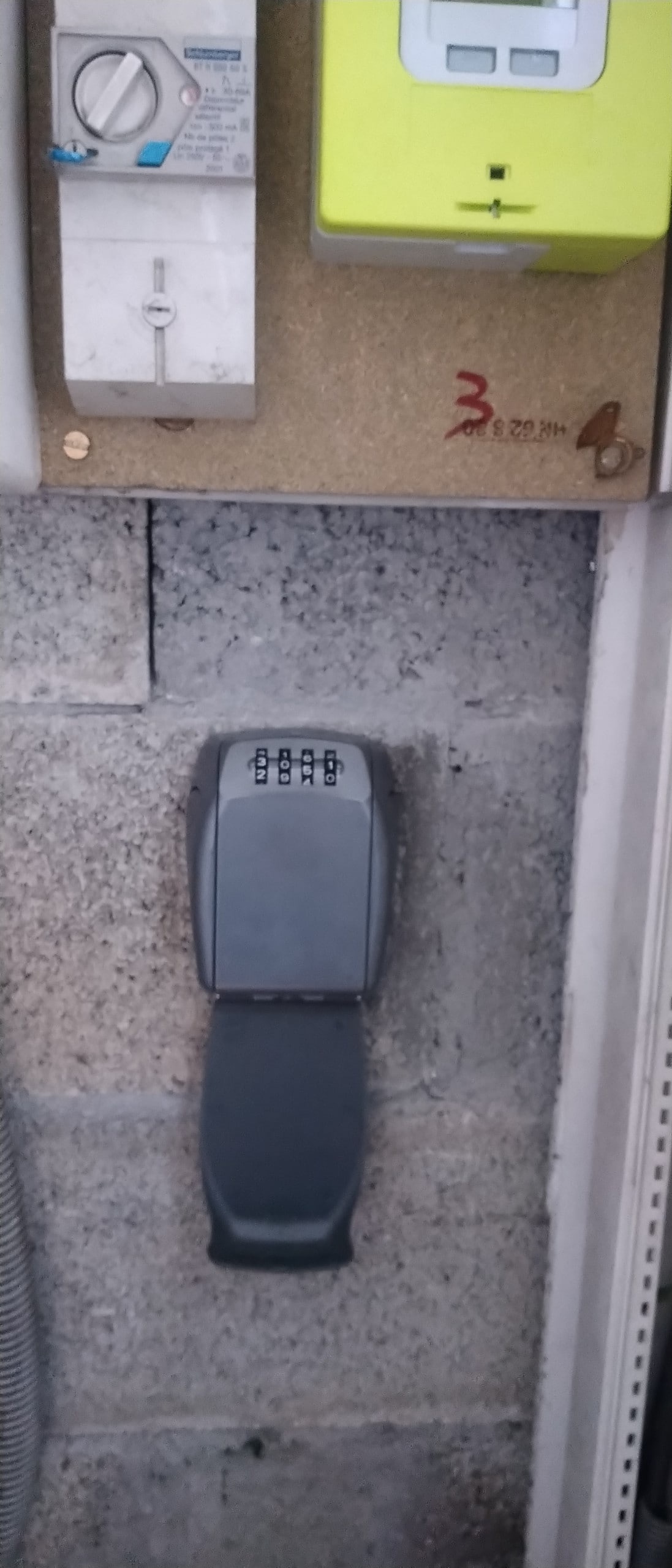 A wall-mounted key lockbox is visible, secured to a textured surface. The lockbox features a keypad for entering a code and is positioned near a small electrical control panel. Nearby, the wall is made of gray concrete blocks.