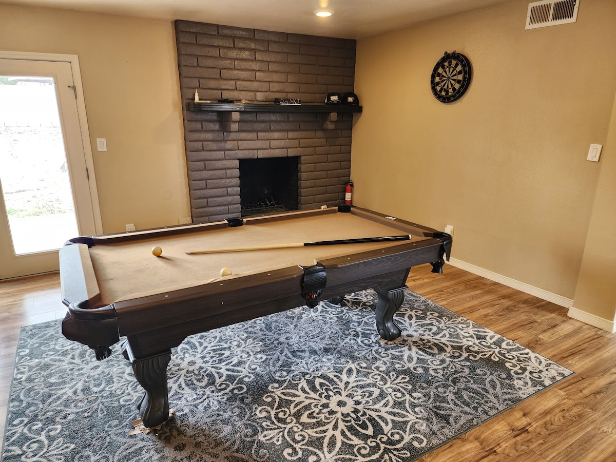 A game room features a pool table positioned on a decorative area rug, with a dark stone fireplace in the background. Natural light enters through a glass door to the side, creating a relaxed environment. A dartboard is mounted on the wall for additional entertainment.