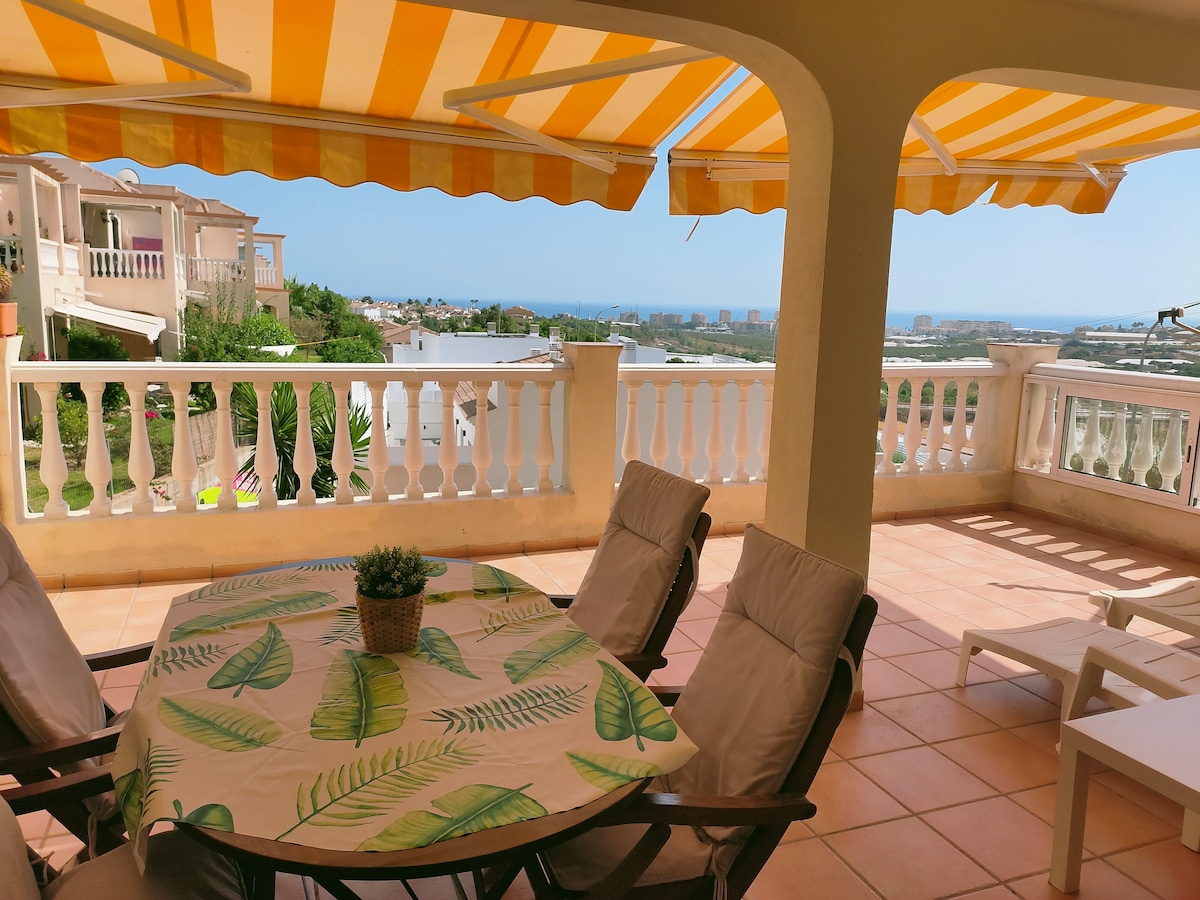 A spacious terrace is featured, showcasing a round table covered with a patterned tablecloth and surrounded by four comfortable chairs. Sun loungers are placed nearby, under a striped awning. The distant view reveals the sea and a nearby town, enhancing the outdoor setting.
