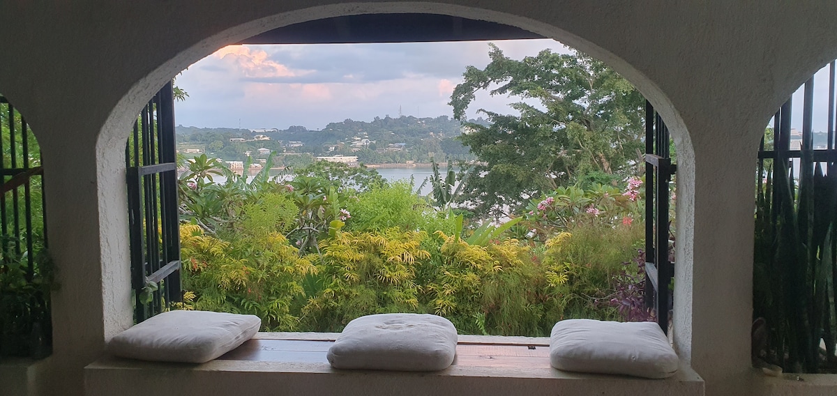 A charming indoor alcove features a wide opening with black grid-like gates, offering a serene view of a lush garden and Port Vila Harbour. Three soft cushions rest on a built-in bench, inviting relaxation while enjoying the natural landscape framed by vibrant greenery.