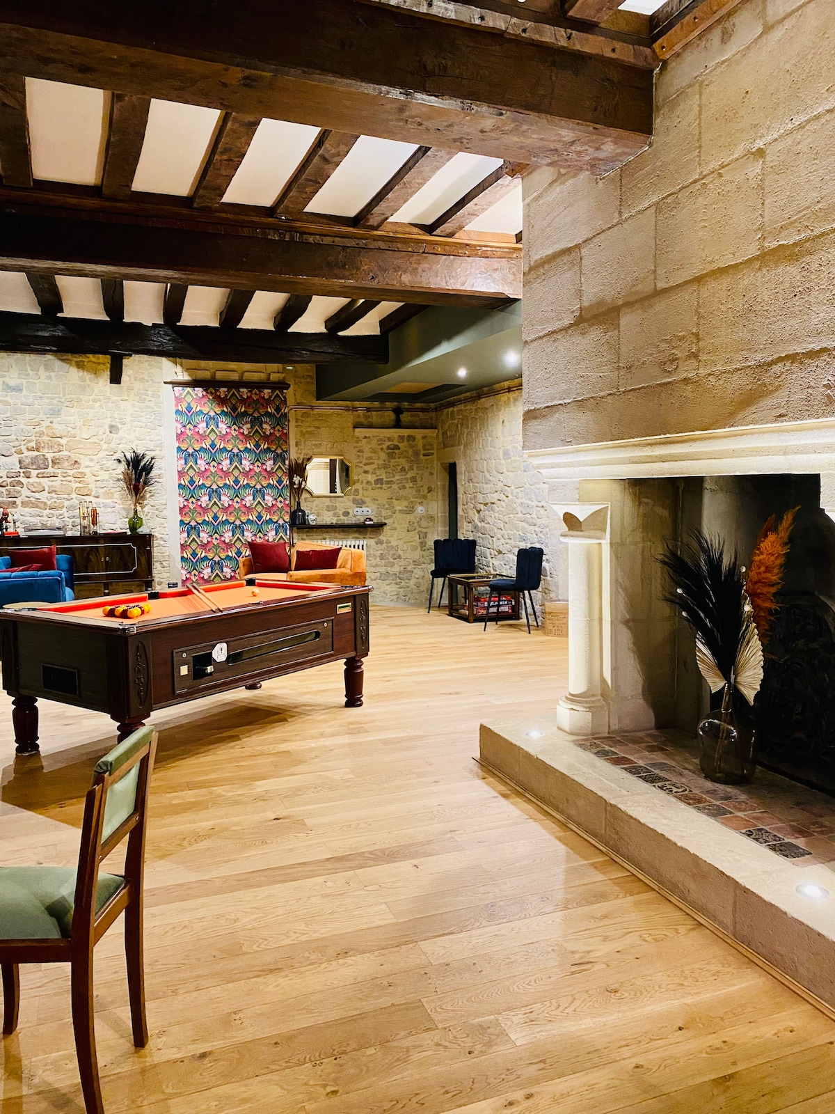 An open living space showcases wooden beams above and a warm-toned wooden floor. A pool table is positioned beside a decorative stone fireplace. Colorful textiles can be seen on the walls, while a small dining table set for four adds to the inviting atmosphere.