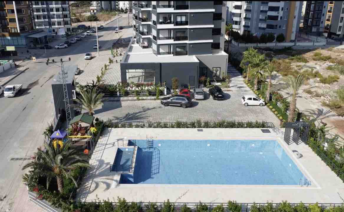 An aerial view captures a modern swimming pool with clear blue water, surrounded by palm trees. The pool area is bordered by greenery and features lounge chairs. Nearby, the modern complex and on-site parking are visible, along with a glimpse of the surrounding area.