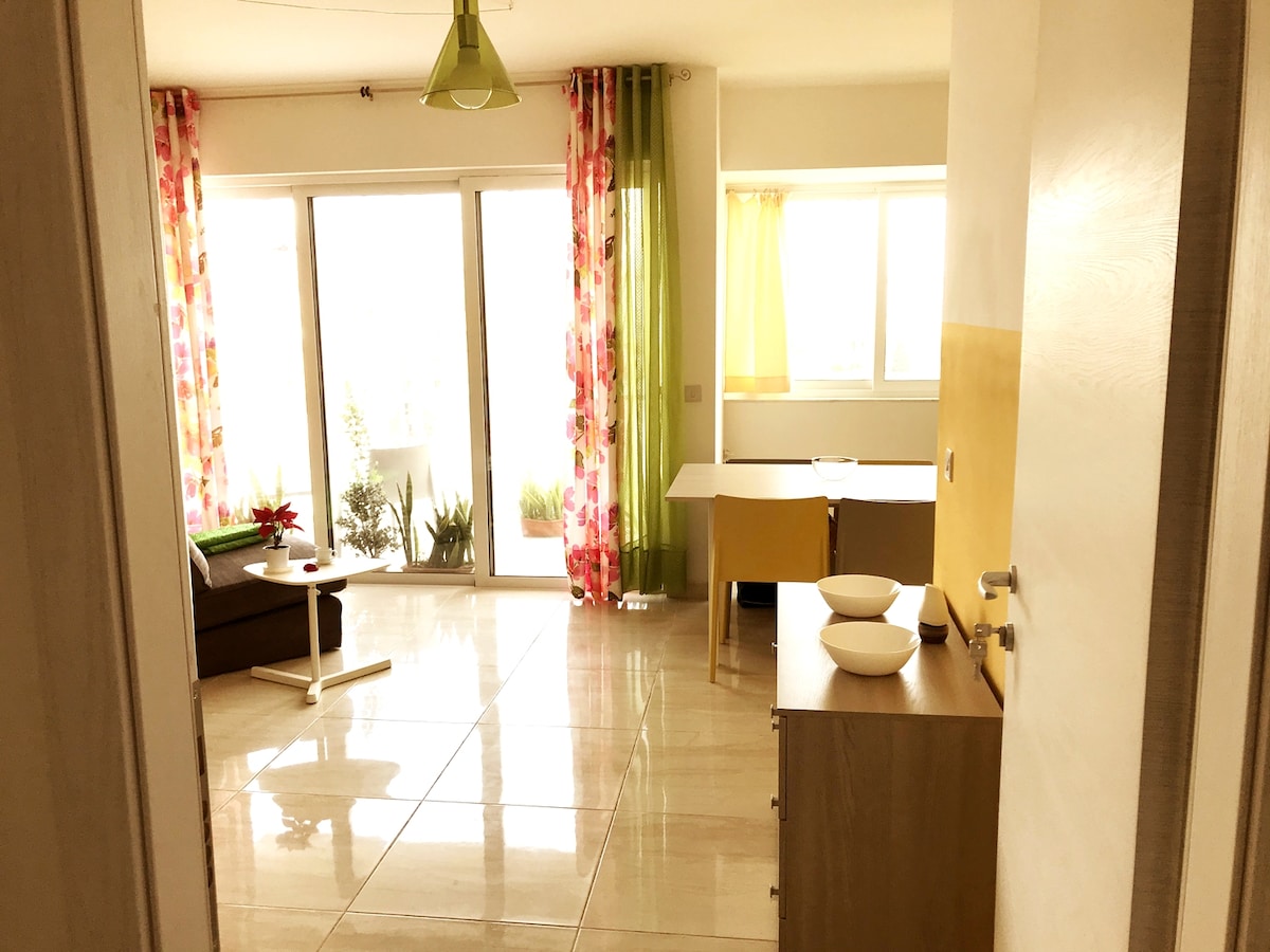 A bright living area is visible, featuring large glass doors that provide access to natural light and a view of greenery. Colorful curtains frame the windows, while a modern sofa and dining table in light hues create a relaxed ambiance.