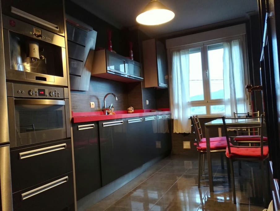 Cocina moderna equipada con vitrocerámica, frigorífico, congelador, horno doble función microondas, lavadora, lavavajillas, armarios, mesa de cristal y cuatro sillas.