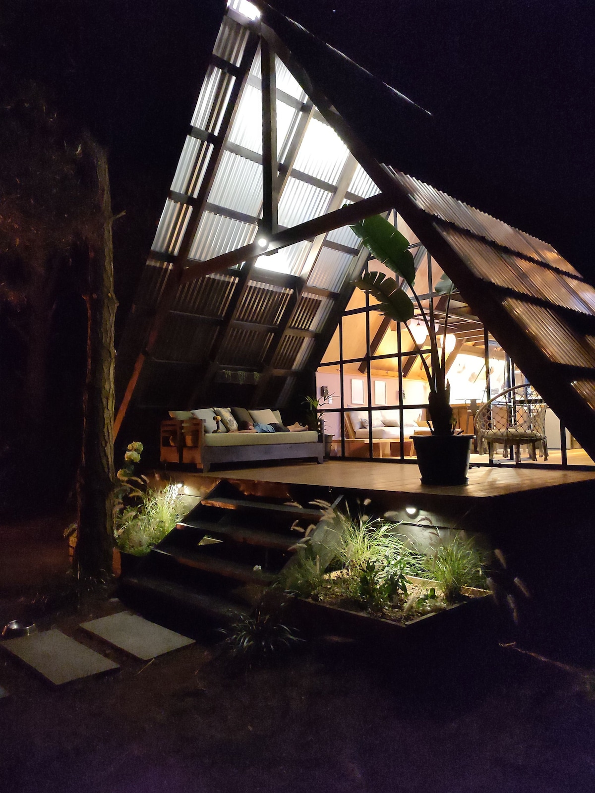 An A-frame house presents a distinctive architectural design under a night sky. Soft lighting highlights the wooden steps leading to a shaded deck adorned with seating. Lush plants are visible, complementing the natural surroundings and enhancing the inviting entrance.