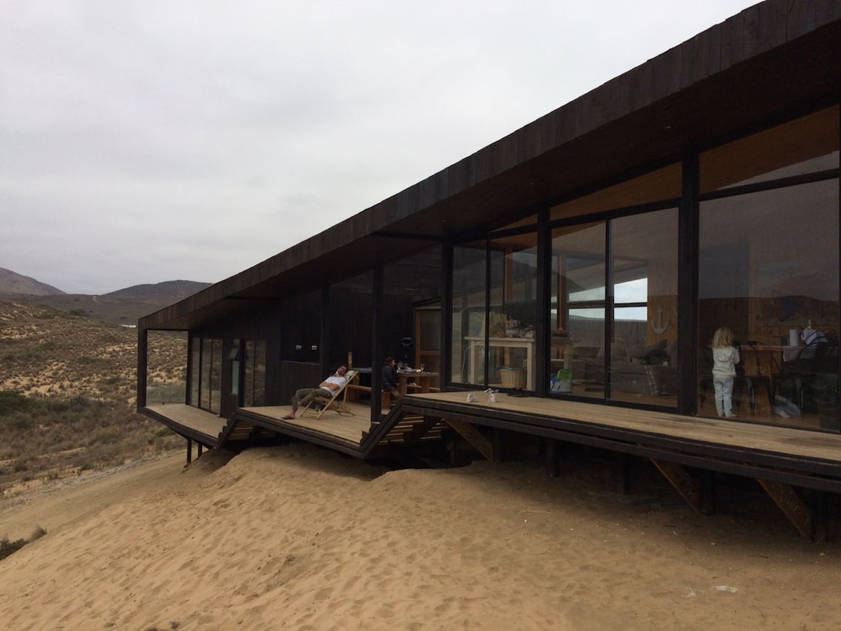 A modern structure sits elevated on a sandy landscape, featuring expansive glass windows that showcase the interior spaces. The design emphasizes a connection with the surrounding natural environment, with a wooden deck extending from the building.