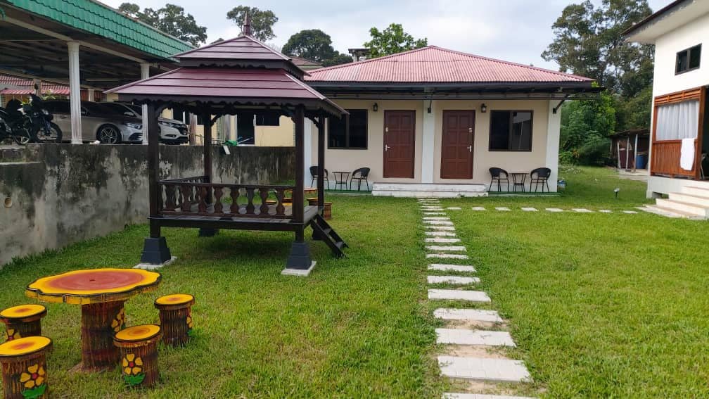 A gazebo with a traditional design is situated in a grassy courtyard, surrounded by stone path tiles. Two doors lead to adjacent units, while colorful tables and stools are placed nearby, inviting relaxation in a natural setting.