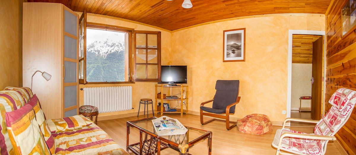 A cozy living room is presented with warm-toned walls and wooden ceilings. A soft seating area includes a patterned couch and a lounge chair. Large windows offer a view of the mountains. A television is mounted beside a small table, creating an inviting space for relaxation.