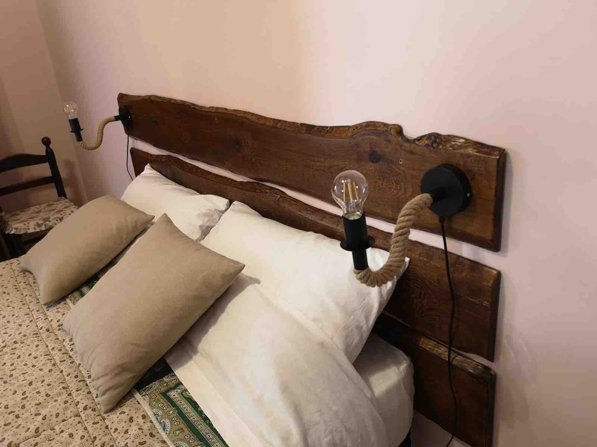 A comfortable sleeping area is featured with a wooden headboard and two wall-mounted lamps. Soft pillows are arranged on the spacious bed, complemented by a patterned bedspread. Warm lighting is provided by the exposed bulbs on either side of the headboard.