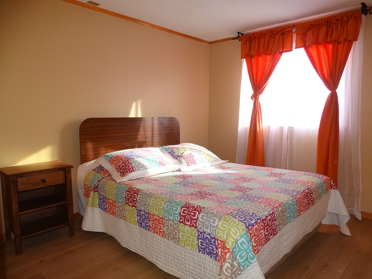 A spacious bedroom features a large quilted bedspread in vibrant colors, complemented by warm wood accents. Natural light floods the room through a bright window, covered by sheer curtains. A small nightstand stands beside the bed, creating a cozy and inviting atmosphere.