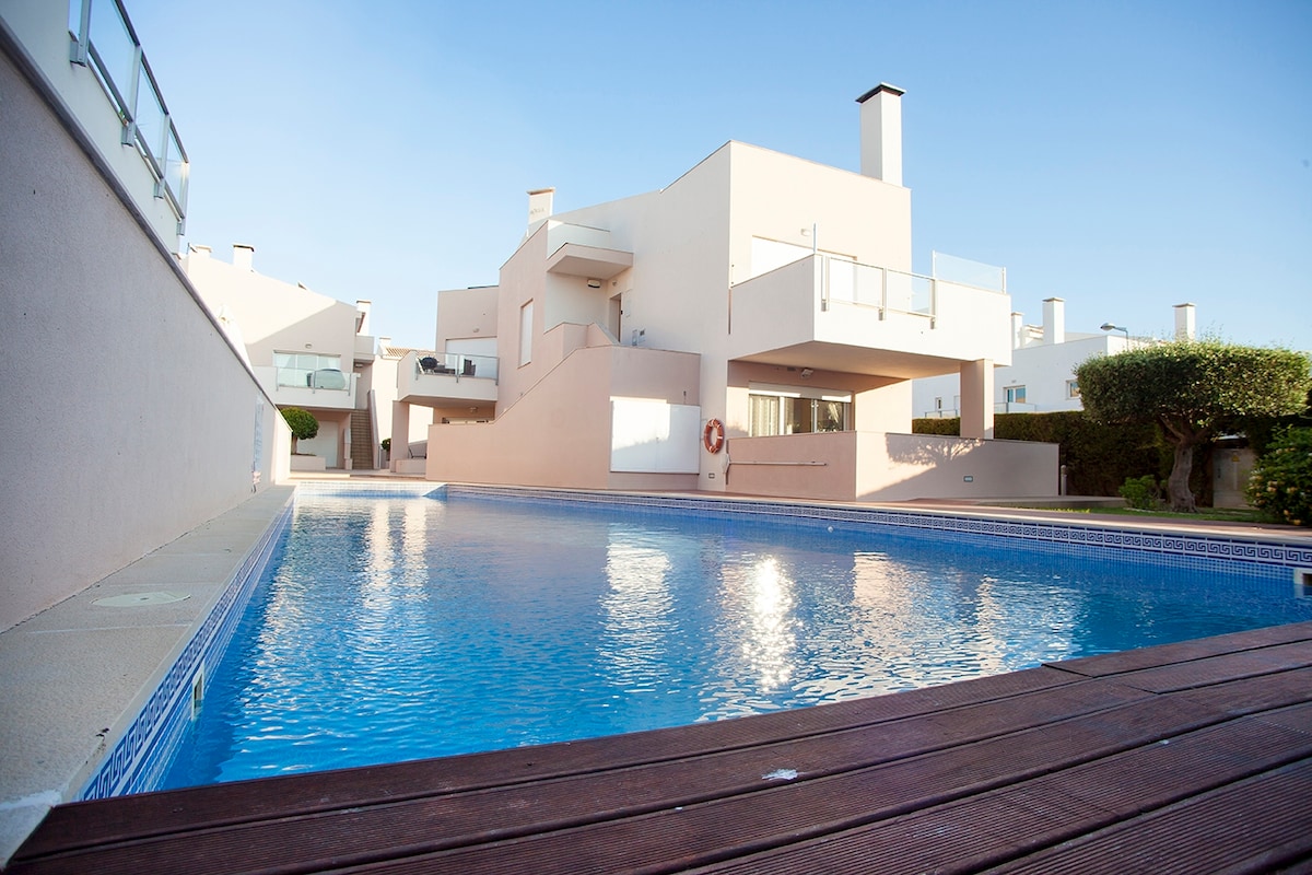 An outdoor swimming pool is framed by modern apartment buildings. The clear water reflects the blue sky, creating a serene ambiance. Wooden decking runs along one side of the pool, providing a space for relaxation or sunbathing.