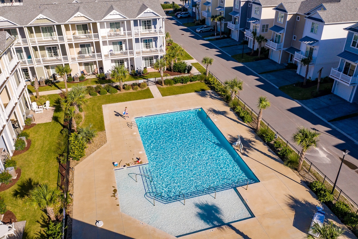 A spacious community pool is set within a well-maintained outdoor area surrounded by lush palm trees. Loungers are positioned poolside, inviting relaxation under the sun. Nearby, residential buildings provide additional context, ensuring a vibrant ambiance.