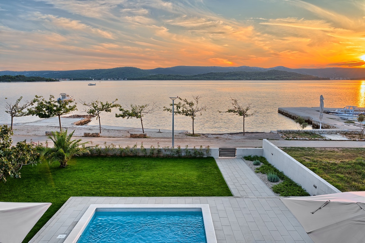 A serene view of the sea is captured at sunset, displaying a tranquil ambiance. The heated pool is surrounded by a spacious deck and a green lawn, while palm trees gently frame the scene. The beach is visible in the distance, enhancing the coastal charm.