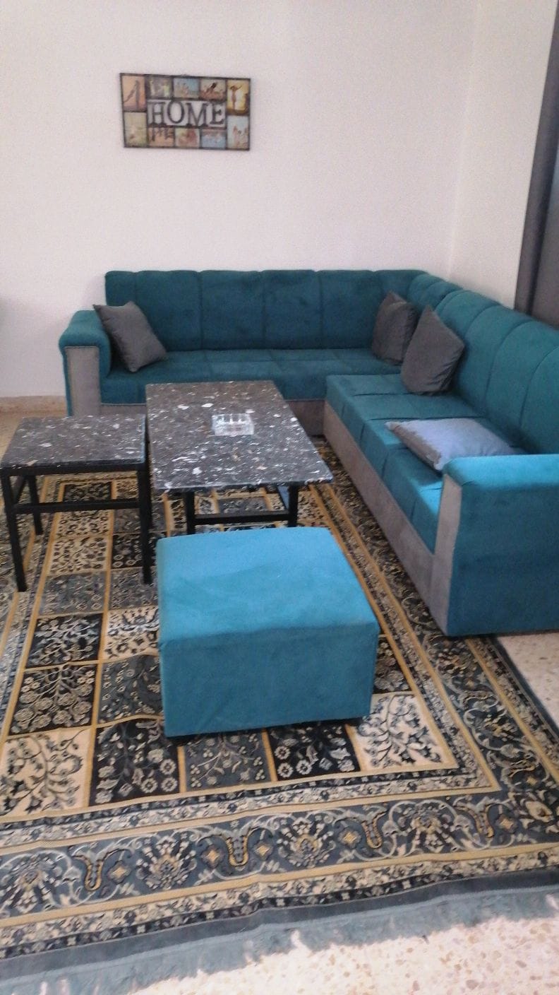 A comfortable seating area is presented with a large blue sectional sofa adorned with cushions. A dark marble coffee table sits centrally, complemented by two smaller tables. An ornate area rug features intricate patterns, enhancing the space's homely feel.