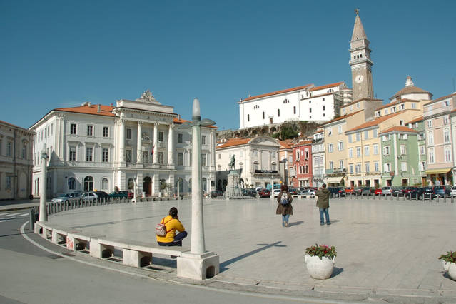 A sunny town square features a spacious plaza surrounded by traditional and colorful buildings. A clock tower rises prominently in the background. People are seen leisurely exploring the area, and decorative plants are placed along the edge of the square, enhancing the inviting ambiance.