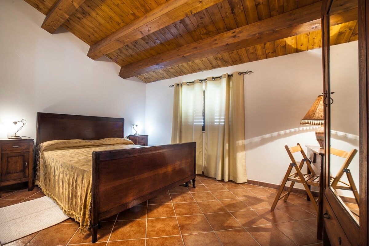A cozy bedroom features a wooden bed with a decorative bedspread, complemented by bedside tables with lamps. Natural light filters through window coverings, casting soft shadows. A small table and chairs are positioned against the wall, and the ceiling showcases rustic wooden beams.