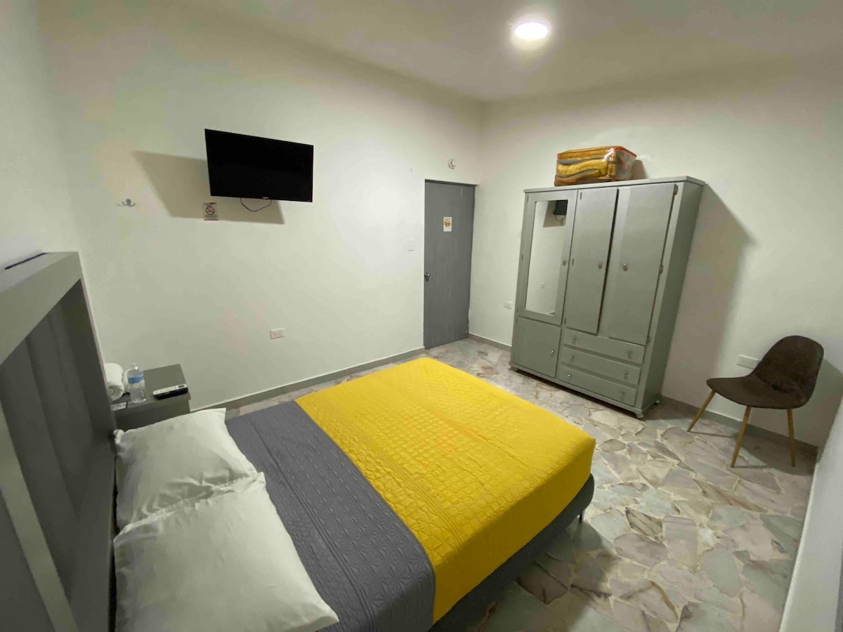 A room is presented with a spacious bed featuring a grey and yellow coverlet. A television is mounted on the wall, and a closet with ample storage space stands nearby. A chair occupies one corner, while the floor displays a mix of light-colored tiles.