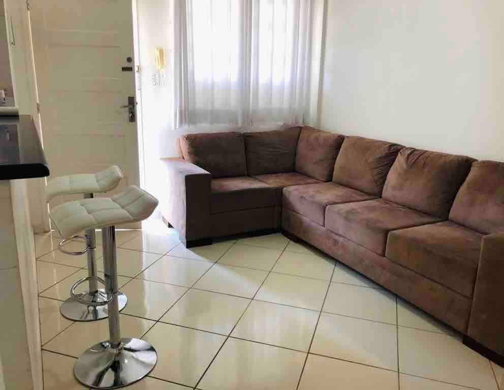 A spacious living area features a large sectional sofa in a neutral tone, providing ample seating. Two high stools are positioned at a counter, with natural light entering through a nearby window, complemented by light-colored curtains.