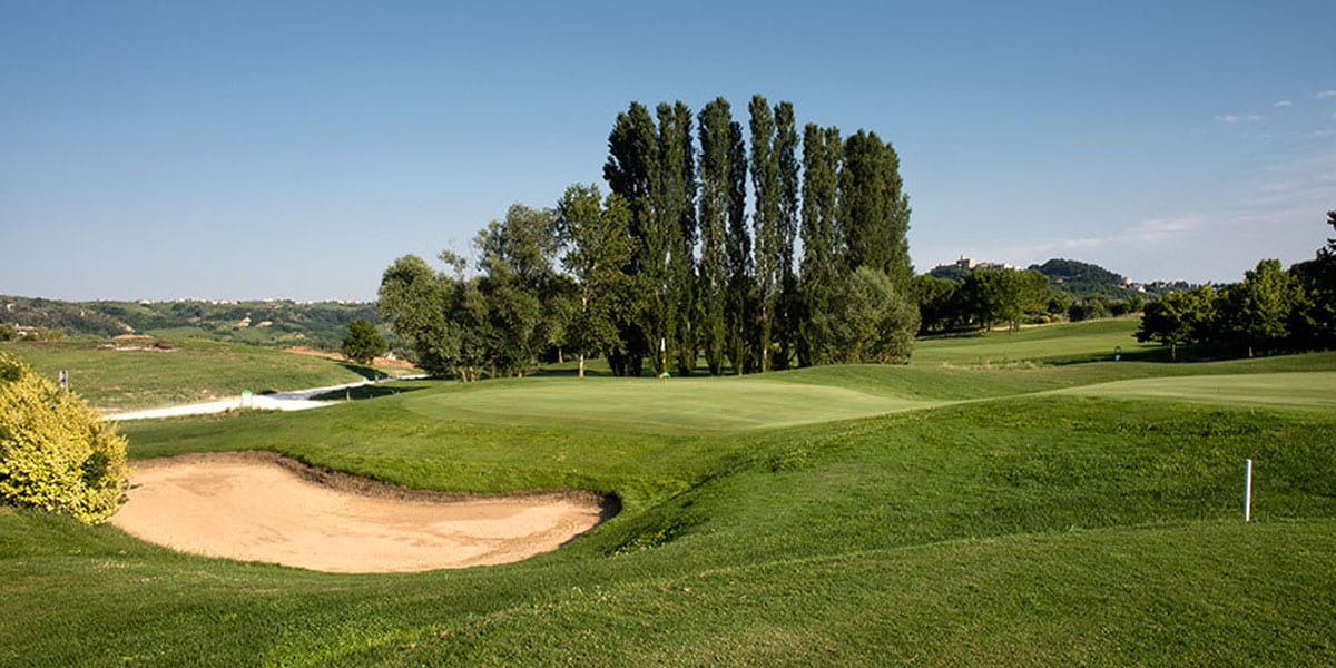 The image captures a lush green golf course, featuring manicured fairways and a sand trap. Tall trees line the background, providing a natural border to the scenic landscape. The distant hills can be seen under a clear blue sky, enhancing the tranquil setting.