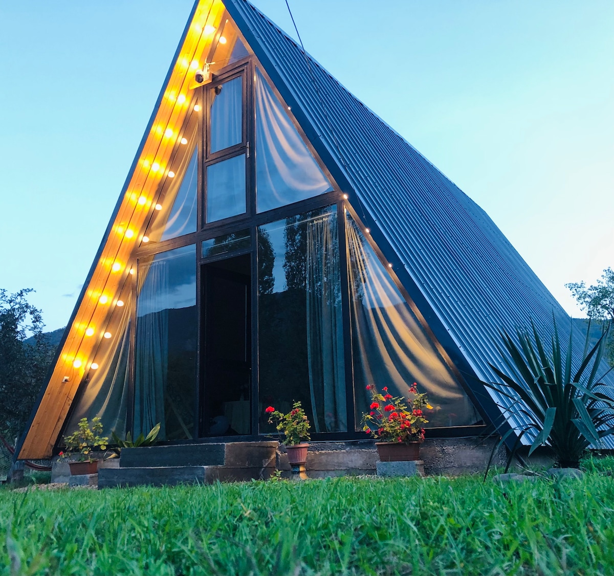 An A-frame cottage is set against a twilight sky, featuring large glass panels that invite natural light. Softly glowing string lights enhance the façade, while potted flowers add a touch of color at the entrance. A grassy area surrounds the structure, creating a serene ambiance.