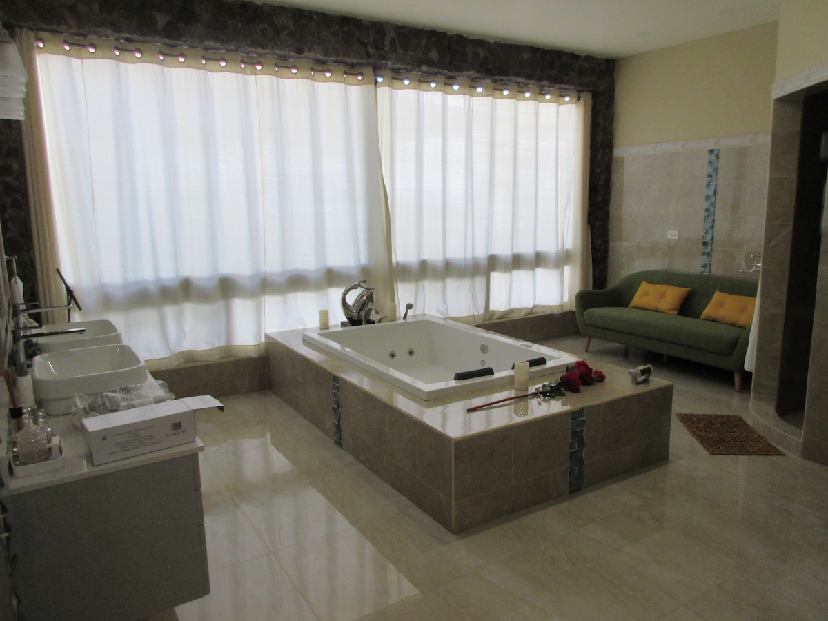 A spacious bathroom features a large jacuzzi tub positioned in the center, surrounded by elegant stone finishes. Natural light streams through sheer curtains, illuminating the area. A comfortable green couch with decorative cushions is placed nearby, adding a relaxed touch to the overall design.